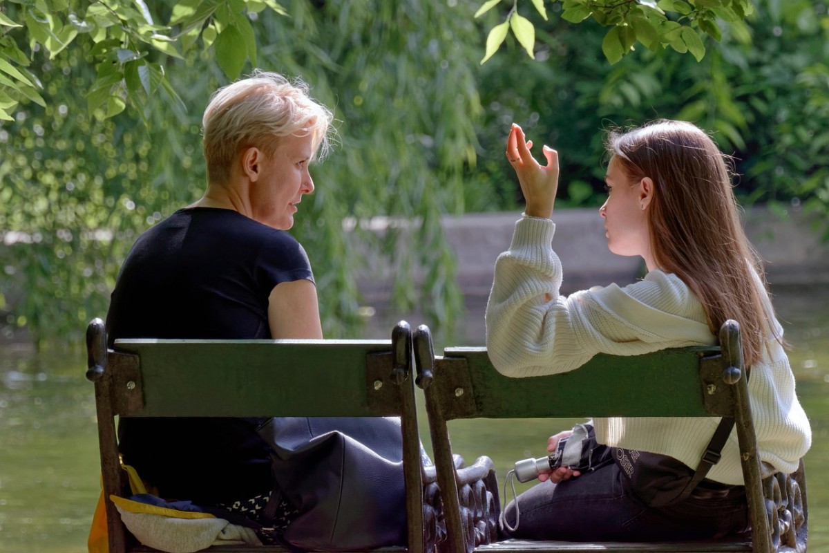 Blond vanem ja tumedapäine noorem naine tiigi taustal pargipingil istumas ja jutustamas